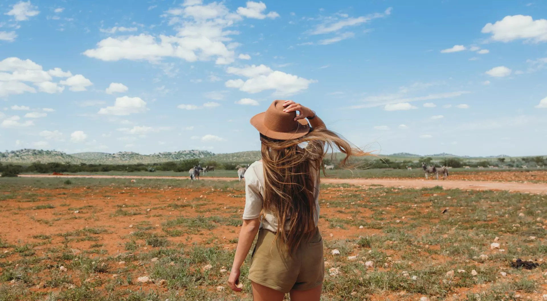 A person with long hair in a hat stands on a grassy plain, looking at a wide sky with scattered clouds.