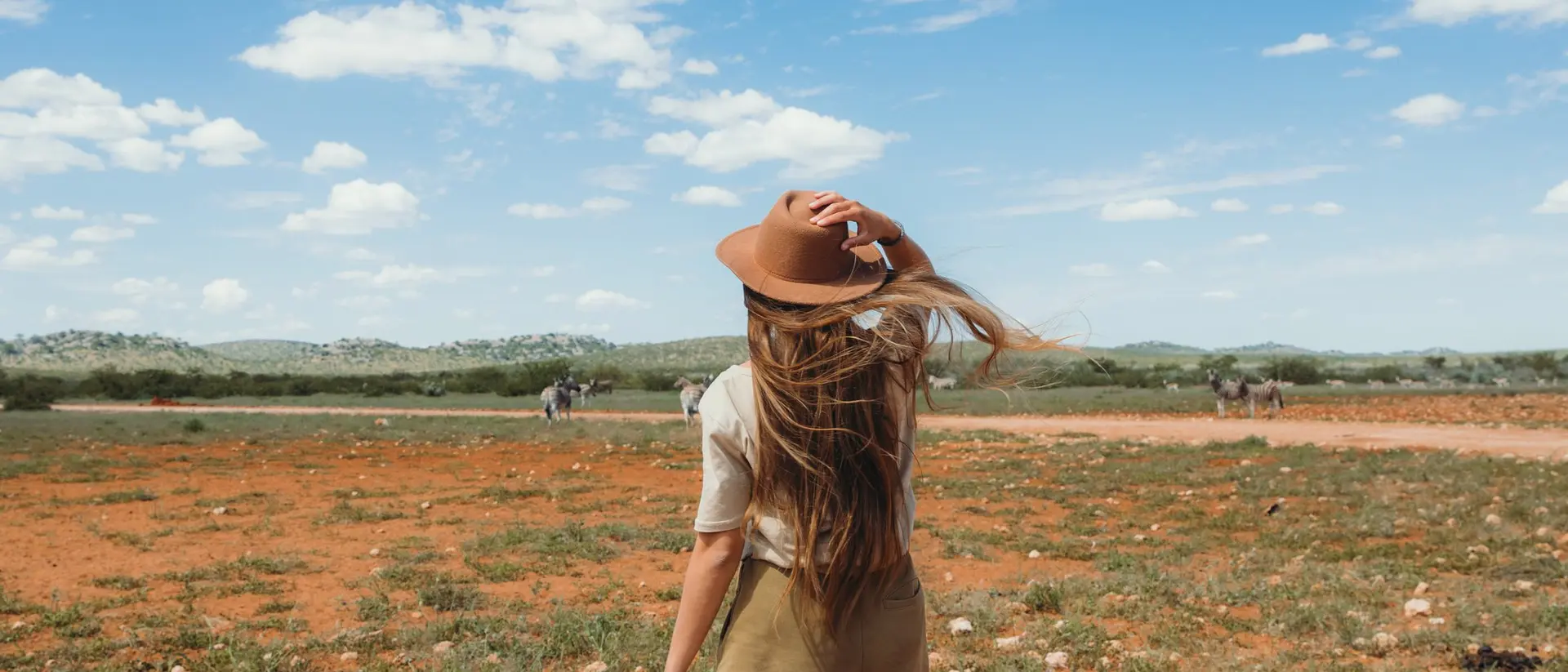 A person with long hair in a hat stands on a grassy plain, looking at a wide sky with scattered clouds.