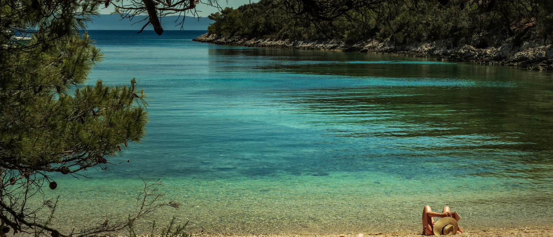A serene beach scene with calm blue water, rocky shores, and a person relaxing on the sand under trees.