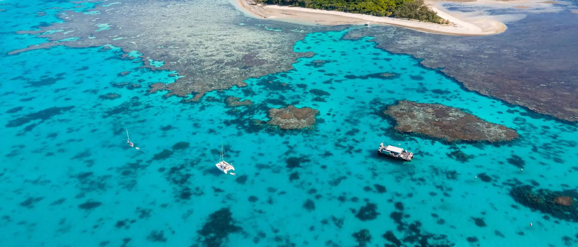Aerial view of a vibrant turquoise sea with coral reefs and a small green island surrounded by sandy shorelines.