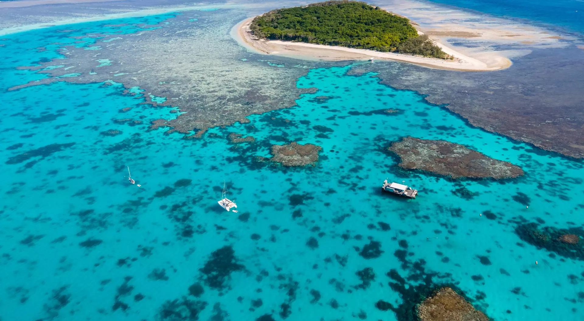 Aerial view of a vibrant turquoise sea with coral reefs and a small green island surrounded by sandy shorelines.