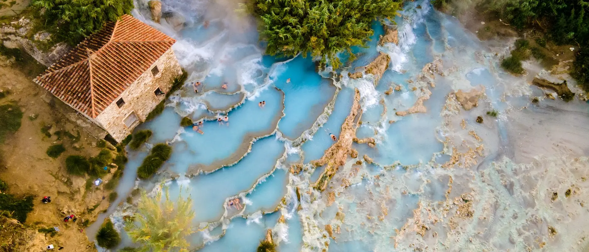 Aerial view of a rustic house near natural hot springs with blue water and lush greenery surrounding the area.
