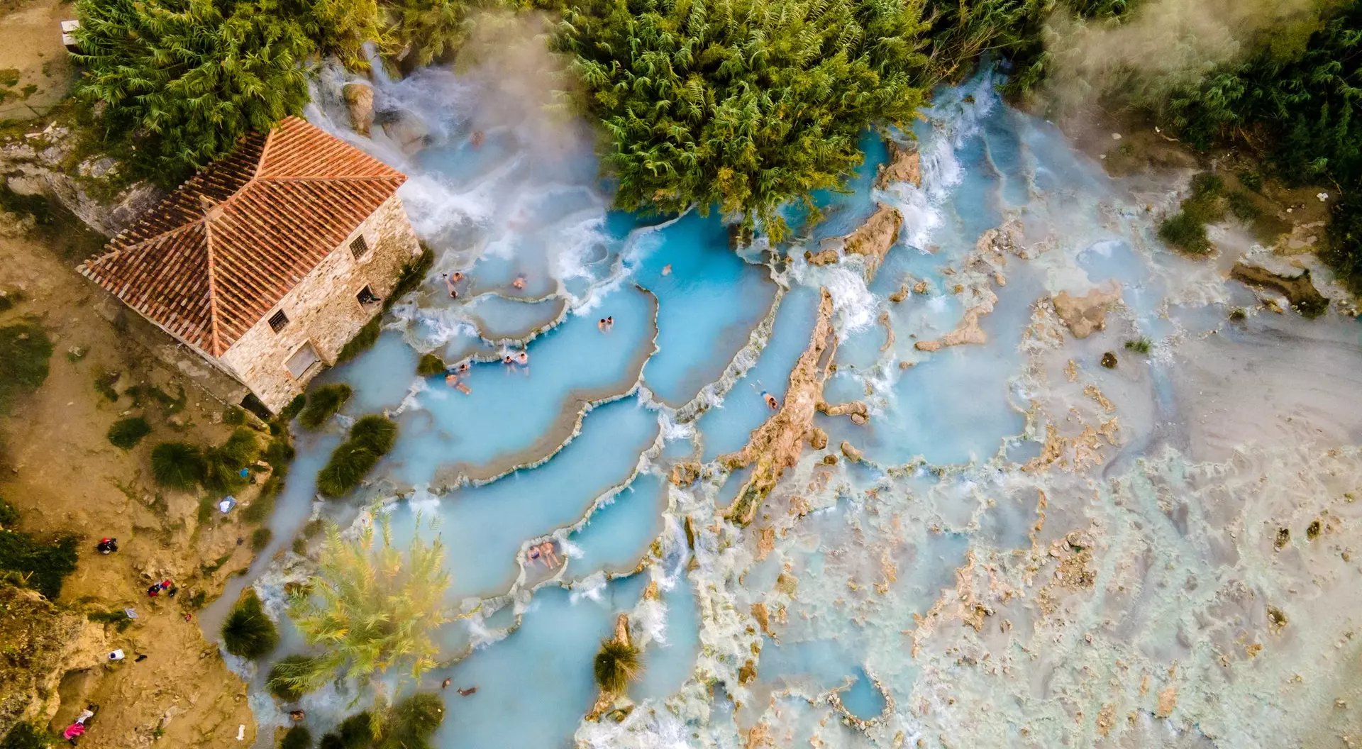 Aerial view of a rustic house near natural hot springs with blue water and lush greenery surrounding the area.