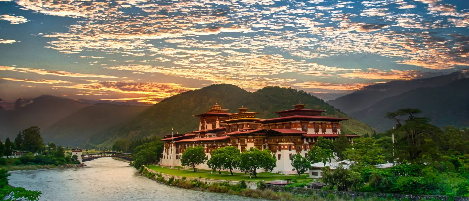A scenic view of a traditional Bhutanese building by a river, surrounded by mountains and a vibrant sky at sunset.