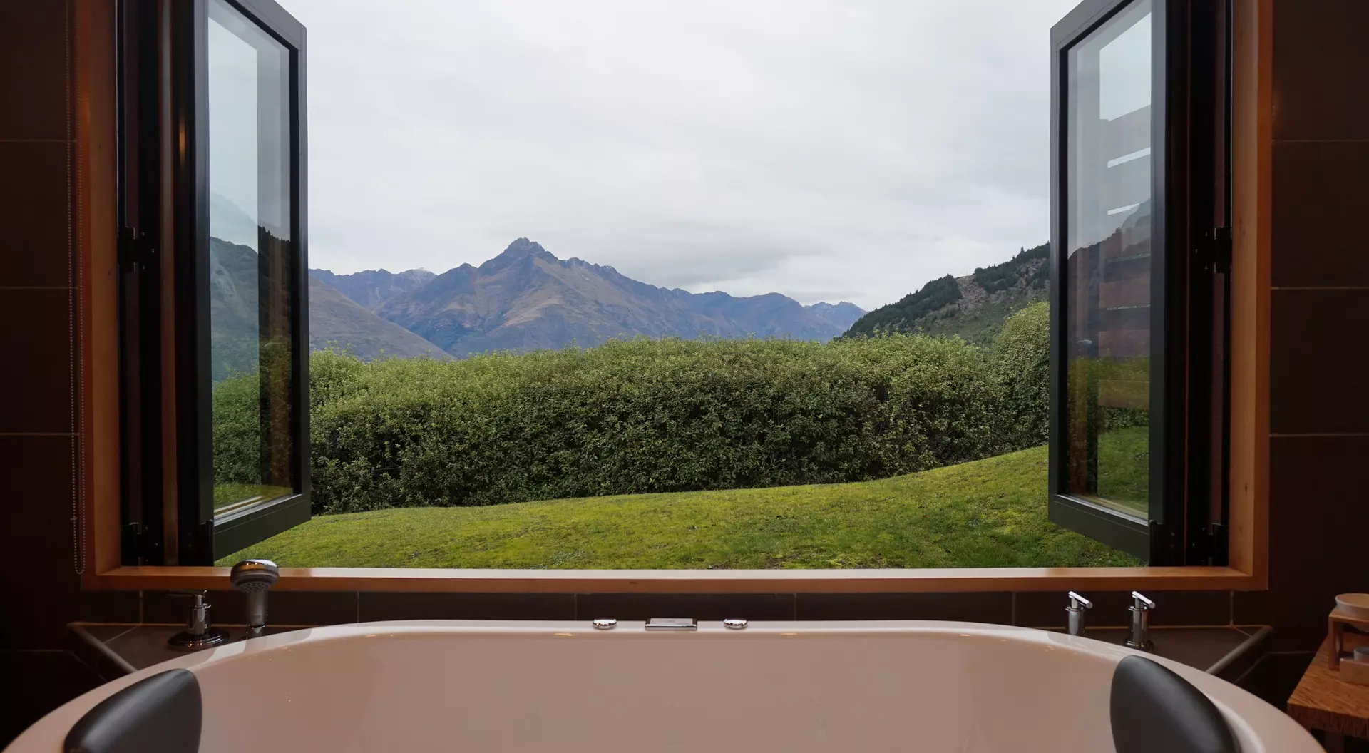 Bathtub in a bathroom with two open windows showing a view of green hills and mountains under a cloudy sky.