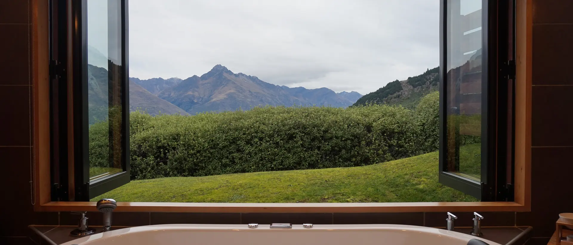 Bathtub in a bathroom with two open windows showing a view of green hills and mountains under a cloudy sky.
