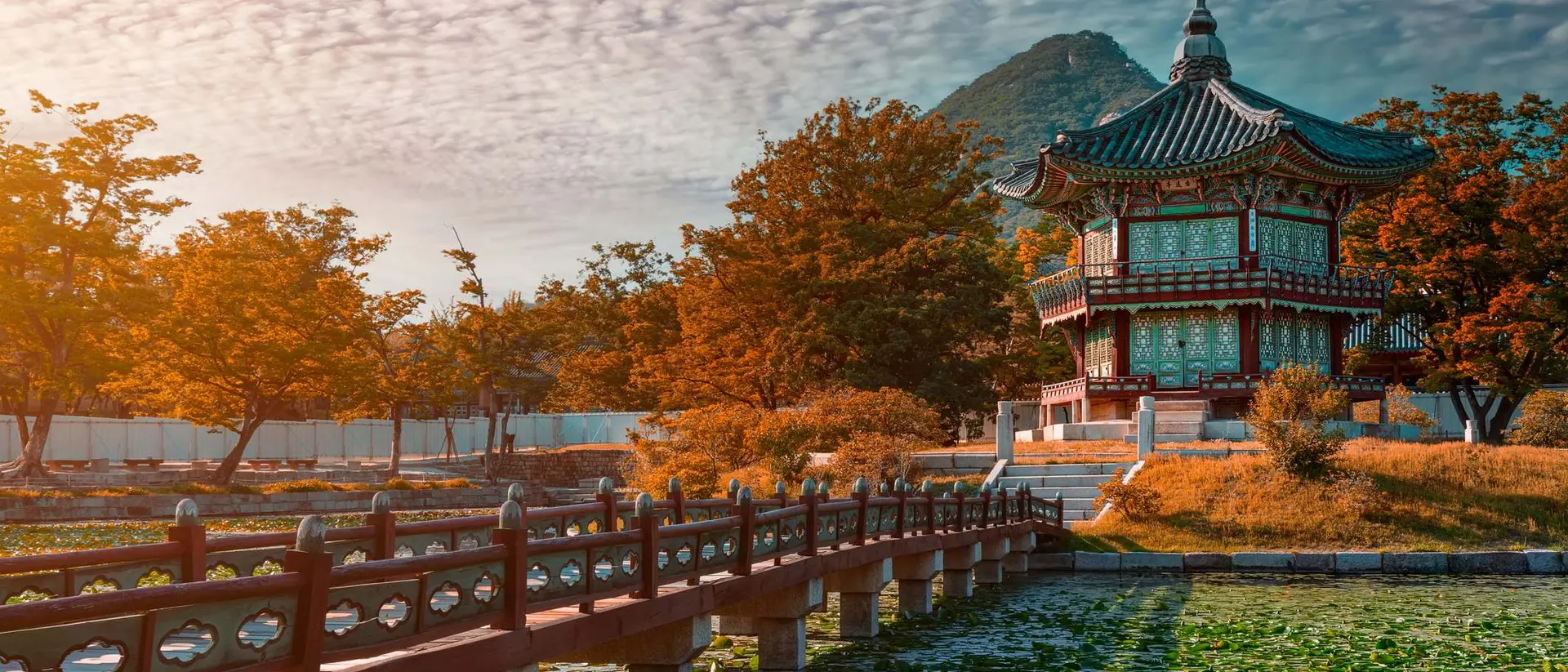 A traditional building with a curved roof beside a pond, surrounded by autumn foliage and mountains under a cloudy sky.