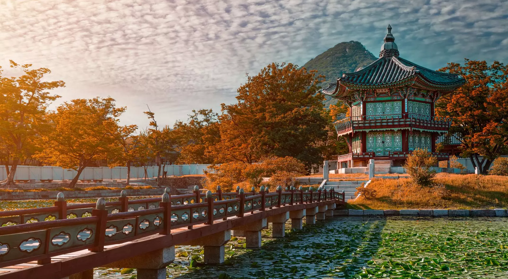 A traditional building with a curved roof beside a pond, surrounded by autumn foliage and mountains under a cloudy sky.