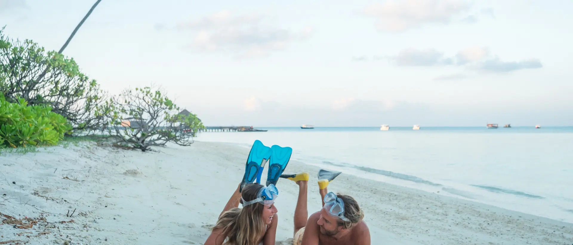 Two people lie on a sandy beach, wearing snorkel fins, with a palm tree and serene ocean in the background.