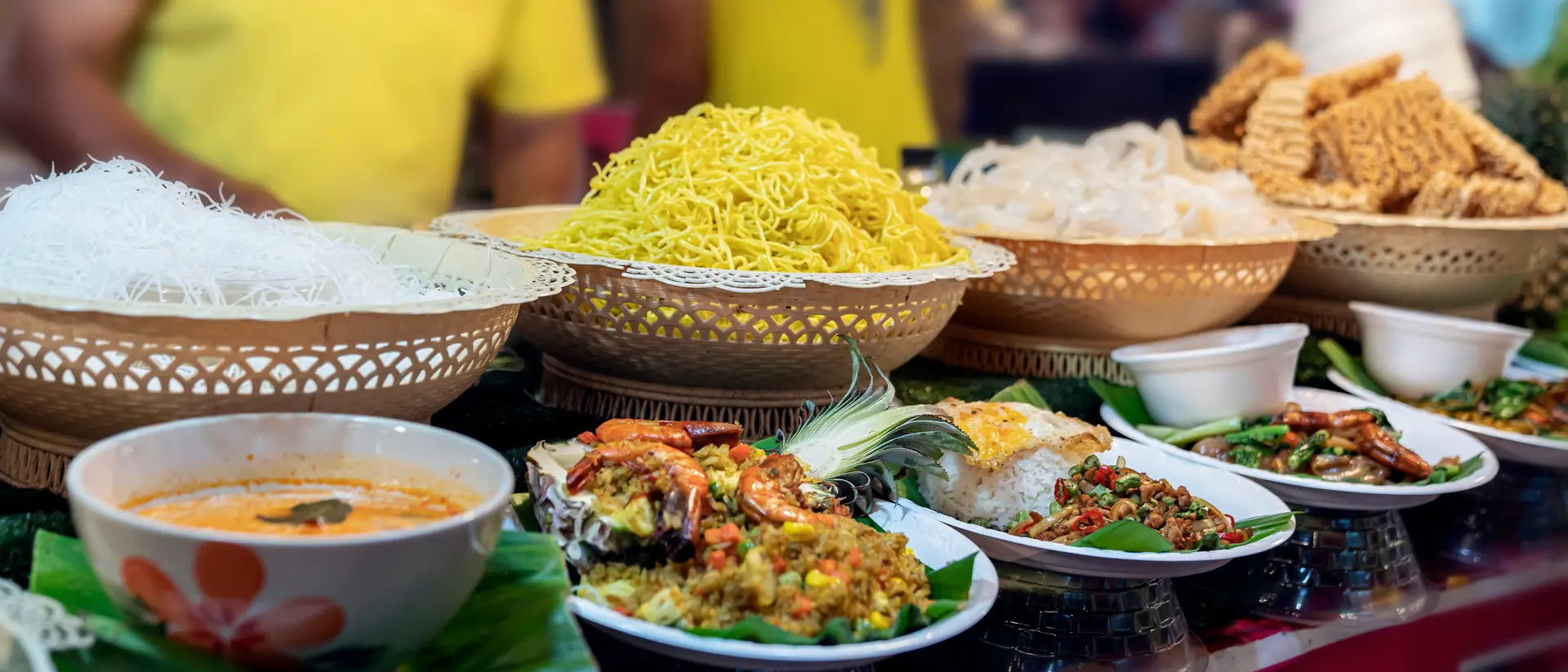 A vibrant food stall featuring various dishes, including noodles, soup, and fried snacks, arranged in baskets and plates.