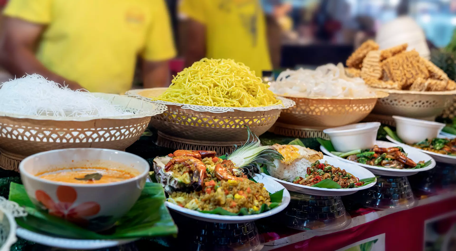 A vibrant food stall featuring various dishes, including noodles, soup, and fried snacks, arranged in baskets and plates.