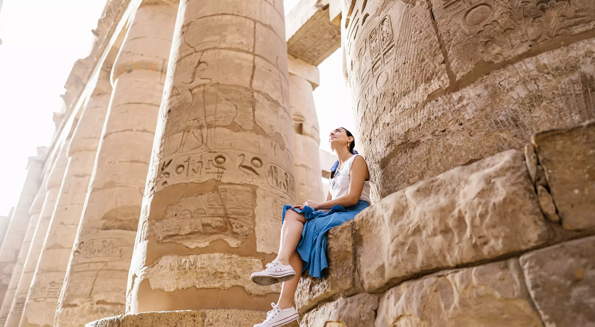Person sitting on a stone block beside tall, ancient columns with hieroglyphics in a sunlit temple.