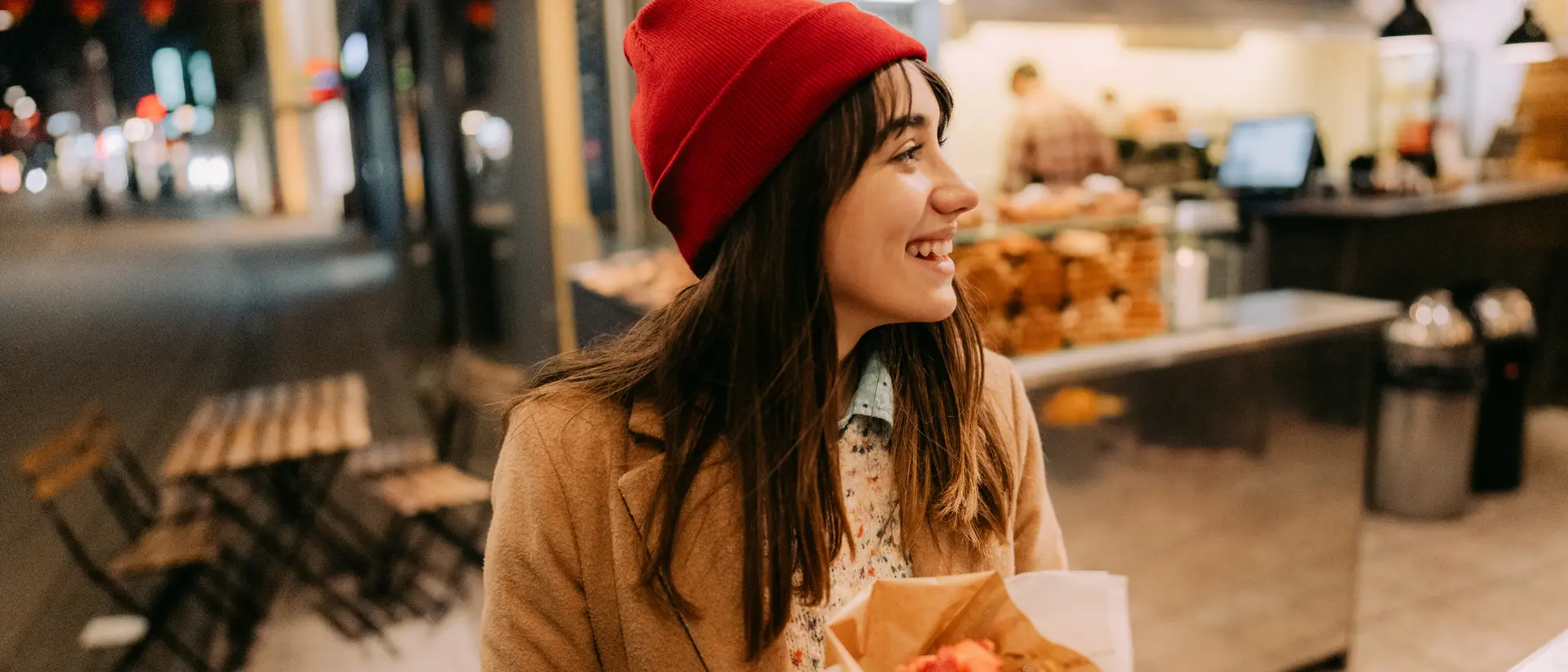 A smiling woman in a red beanie holds a plate of food in front of a cafe at night.
