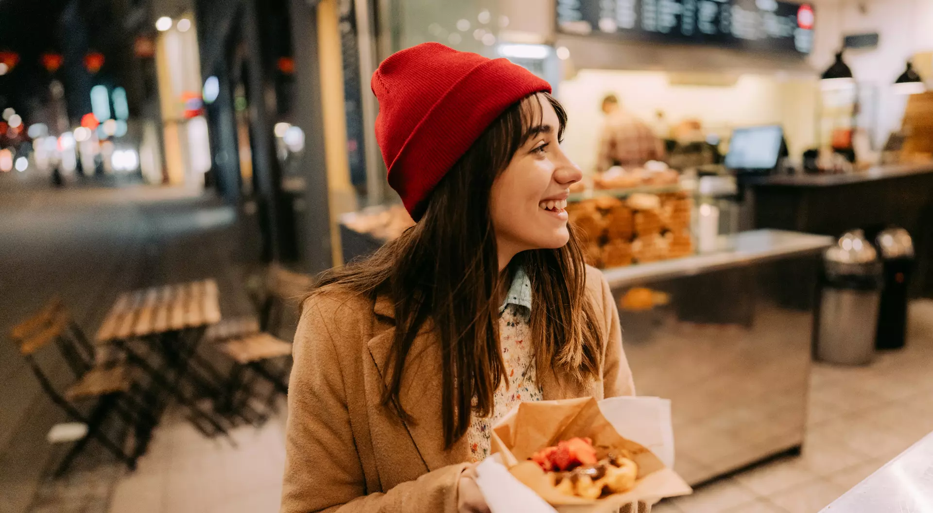 A smiling woman in a red beanie holds a plate of food in front of a cafe at night.