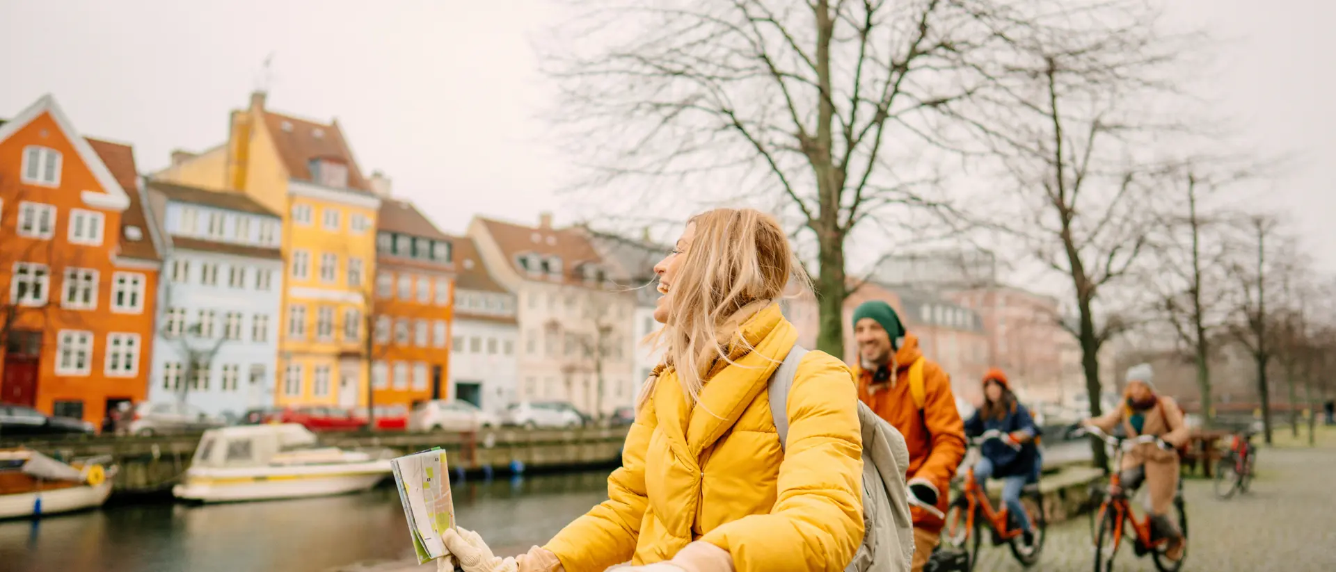 A group of people riding bikes along a canal, with colorful buildings in the background on a cloudy day.