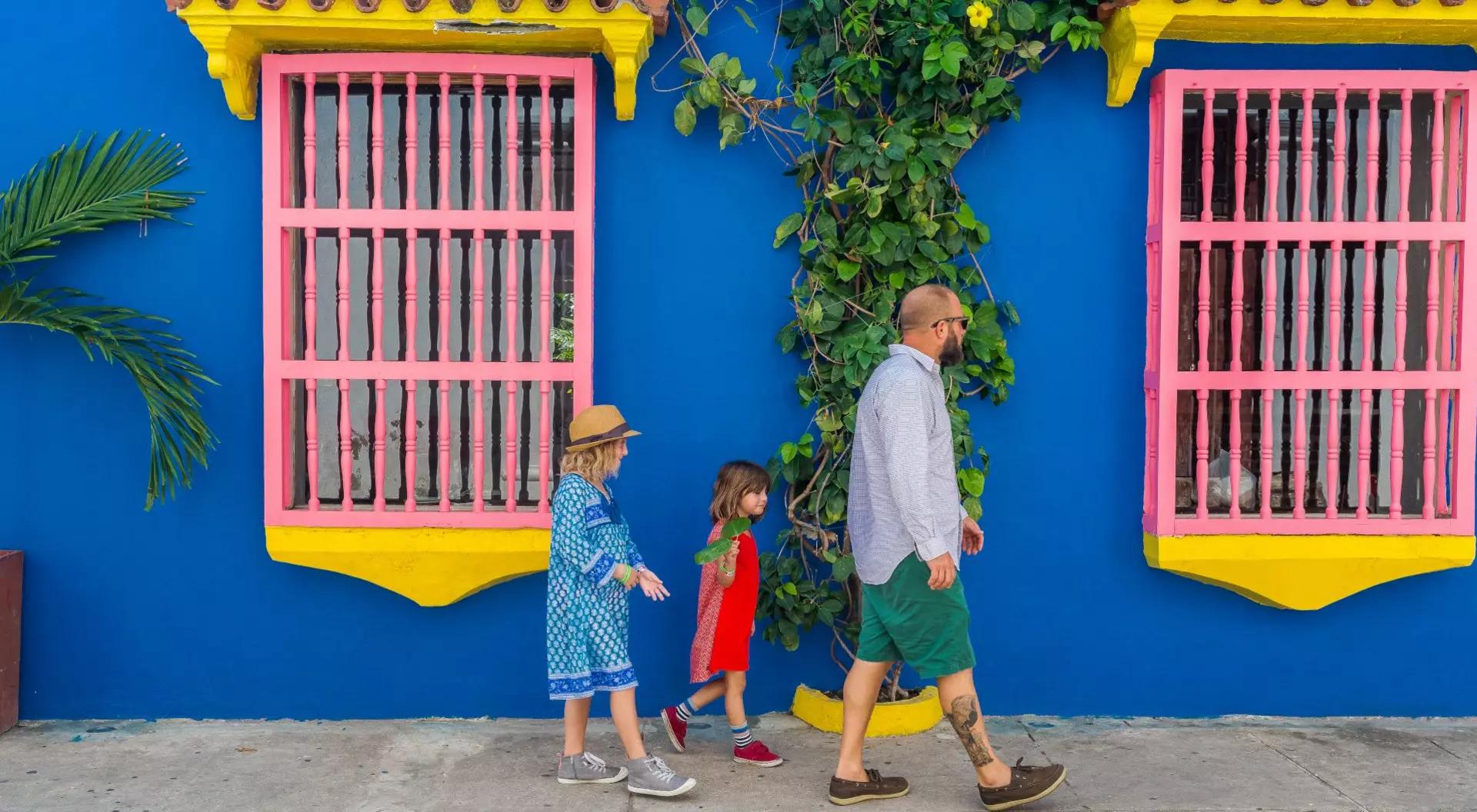 A family of four walks along a bright blue wall with pink windows and greenery, enjoying a sunny day.