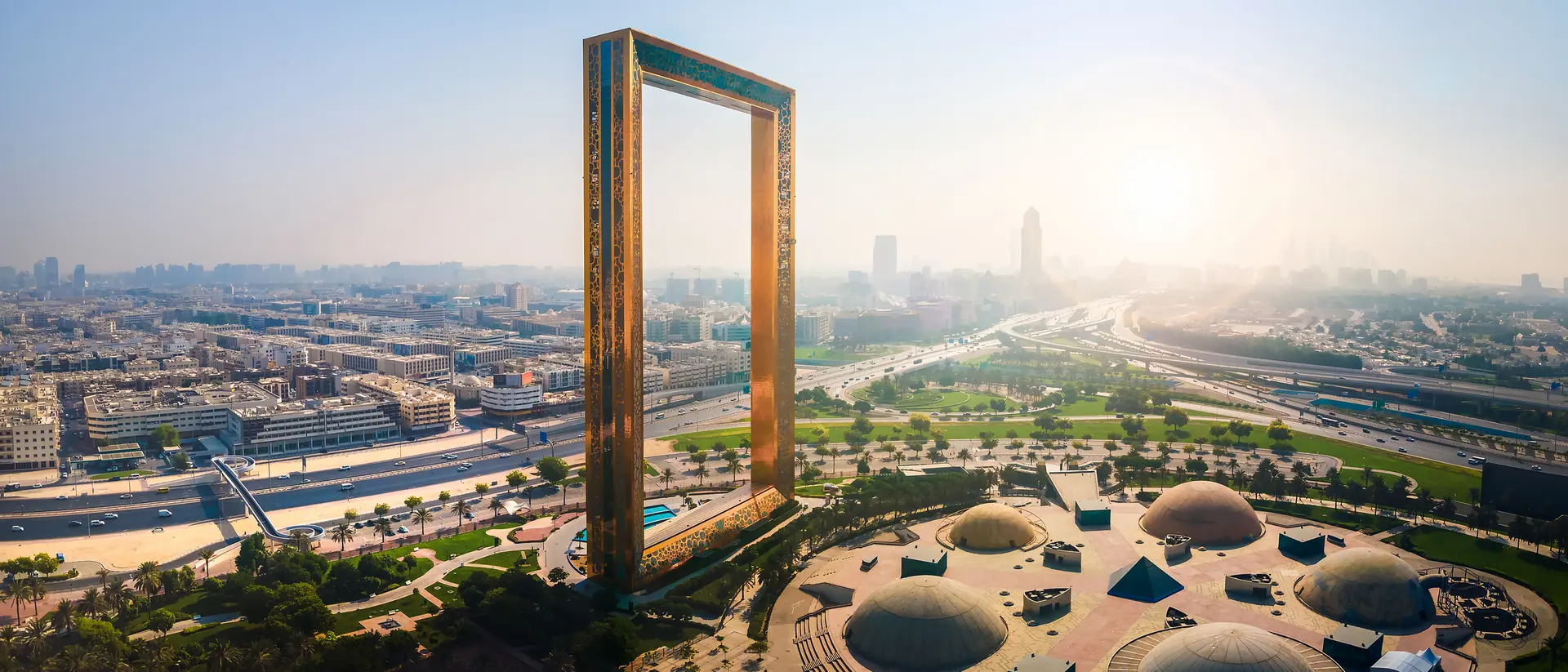 Aerial view of Dubai Frame, a large rectangular structure, with city skyline and green parks in the background.