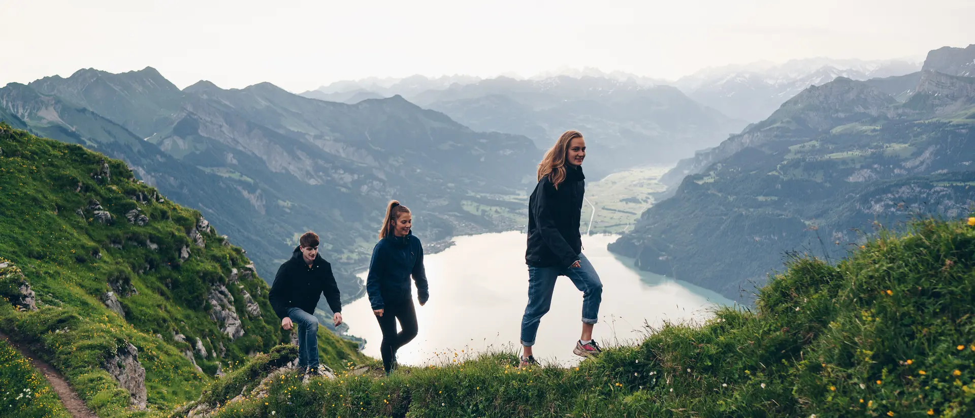 Three people hike along a grassy ridge with a lake and mountains in the background on a cloudy day.