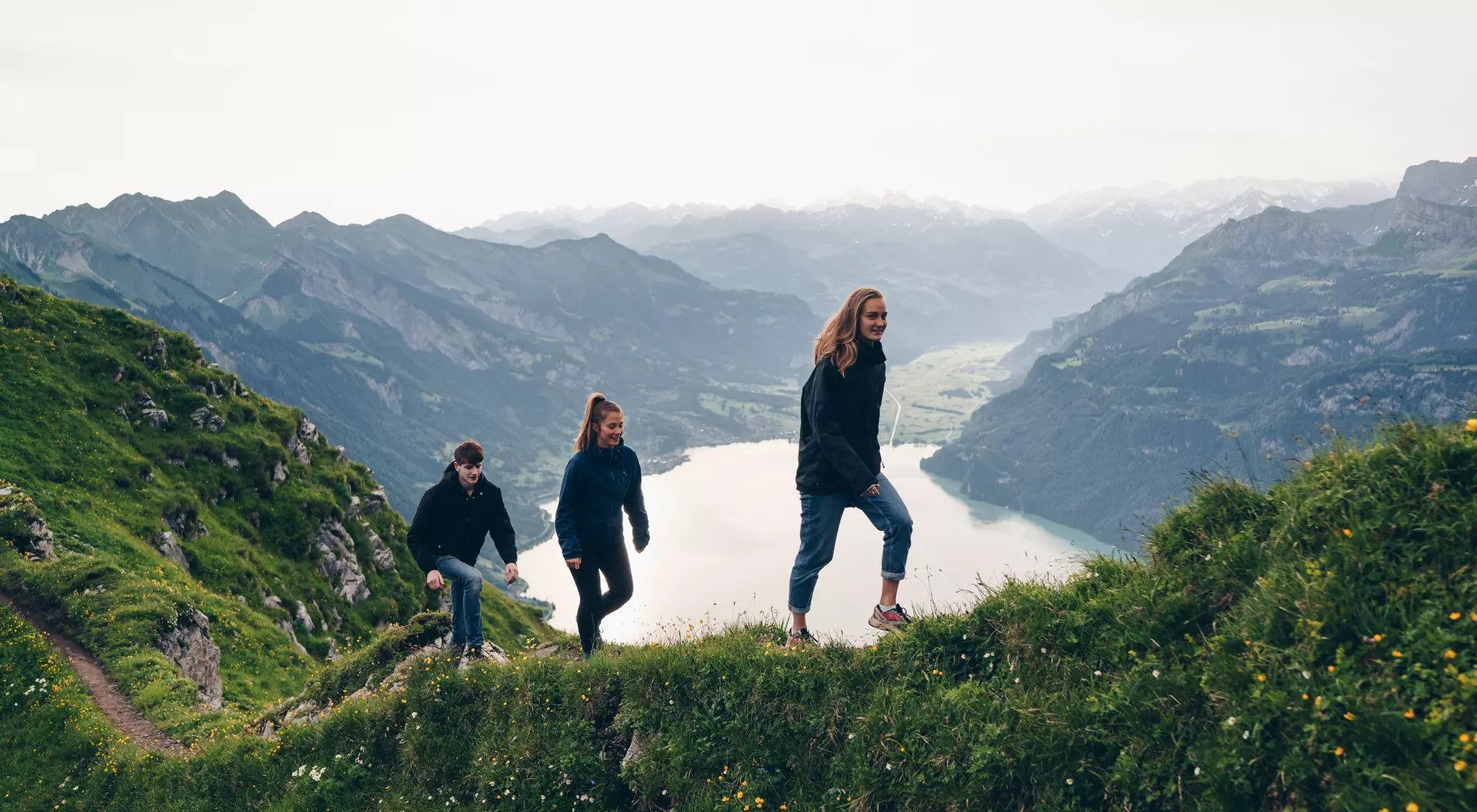 Three people hike along a grassy ridge with a lake and mountains in the background on a cloudy day.