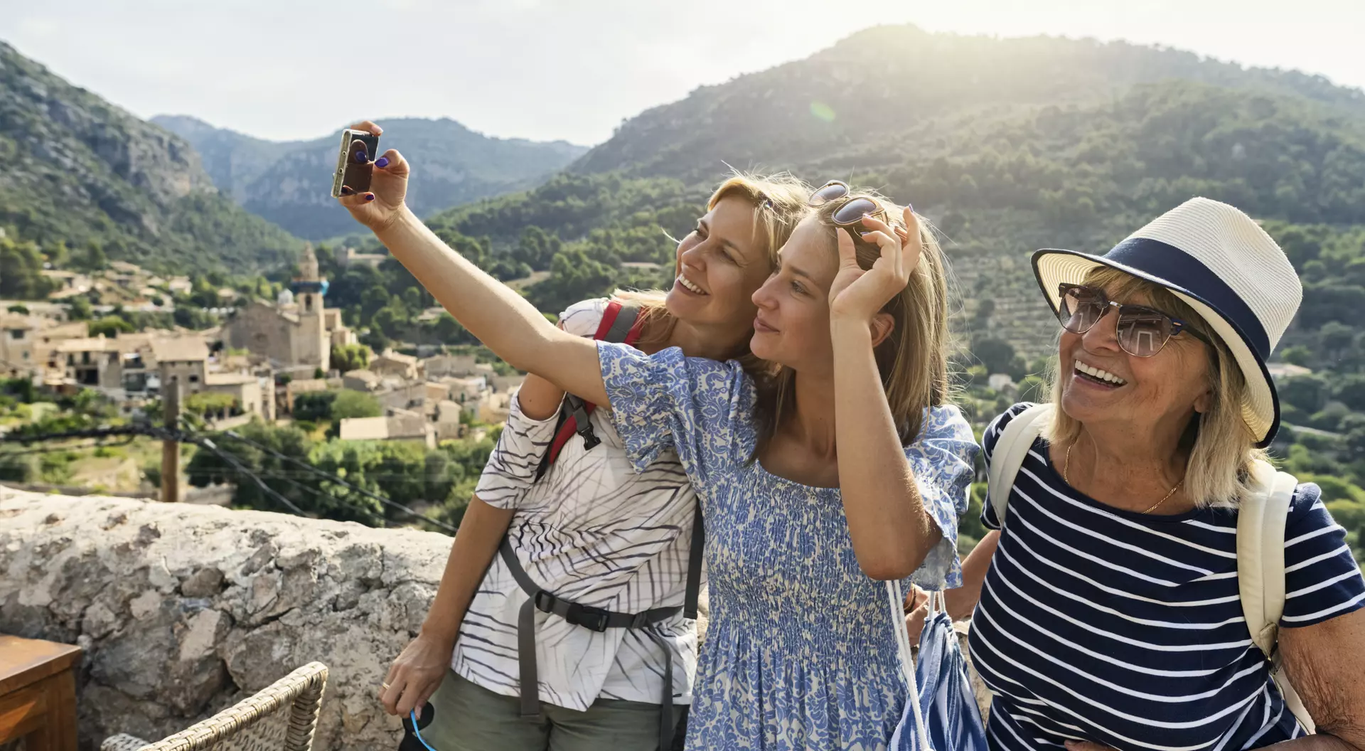 Three women pose for a selfie with mountains and a village in the background, enjoying a sunny day outdoors.