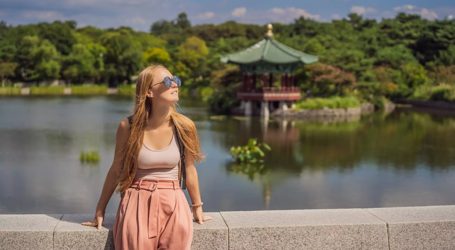 A young woman in sunglasses stands by a lake, smiling with a gazebo and lush greenery in the background.