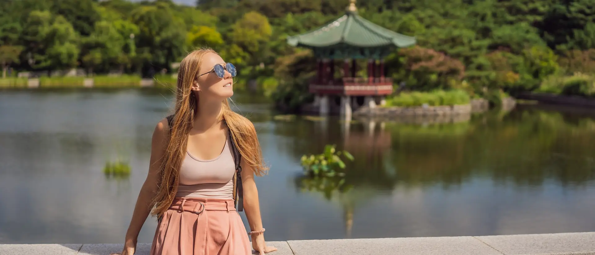 A young woman in sunglasses stands by a lake, smiling with a gazebo and lush greenery in the background.