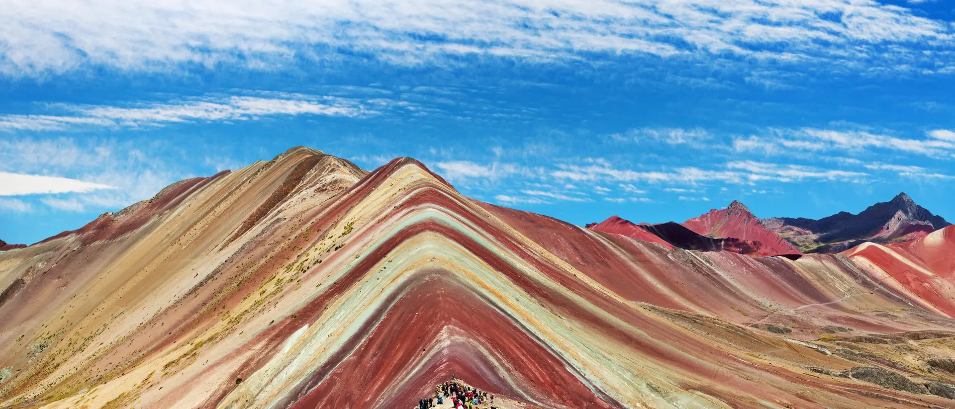 Colorful striped mountain landscape with hikers beneath a blue sky and wispy clouds.