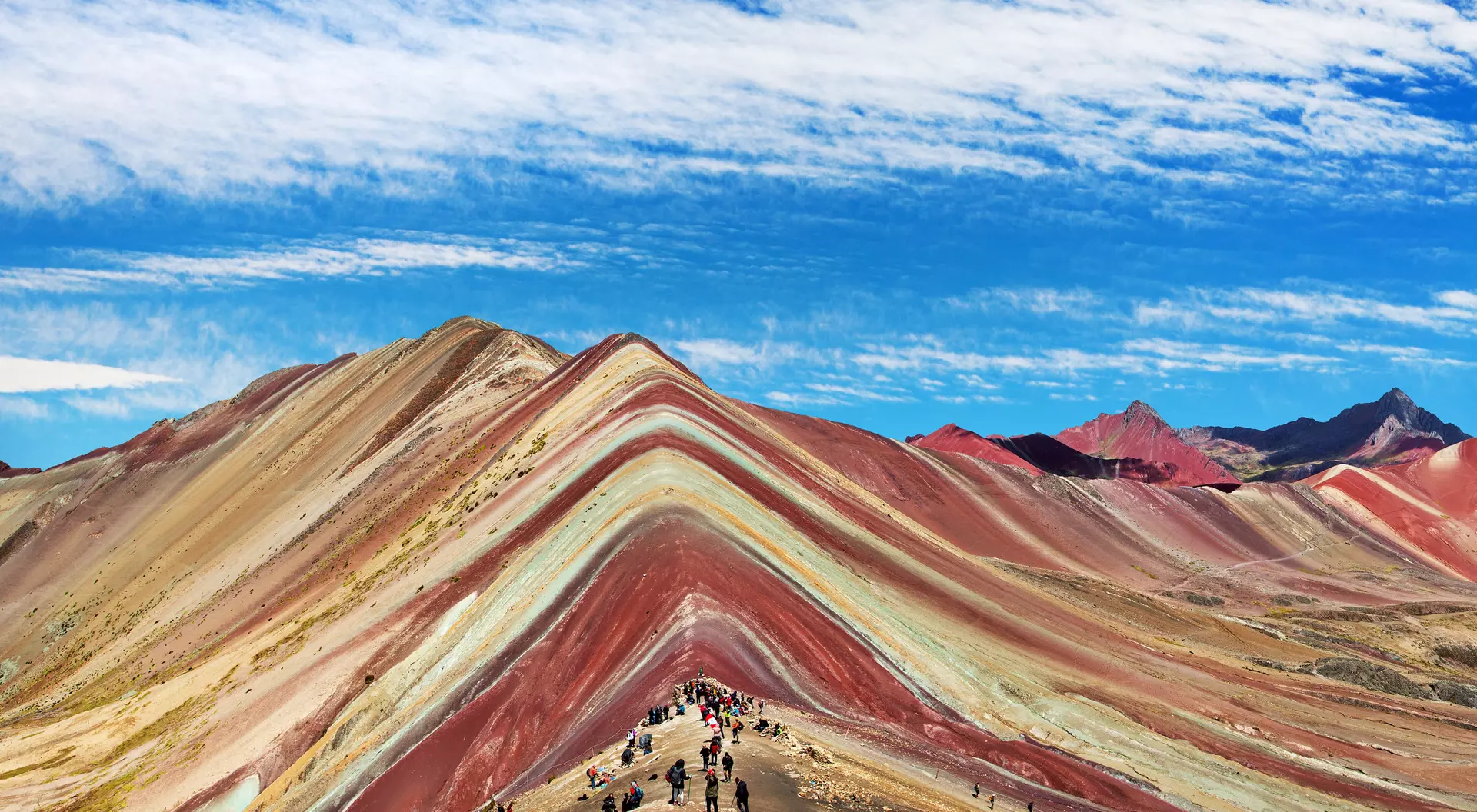 Colorful striped mountain landscape with hikers beneath a blue sky and wispy clouds.