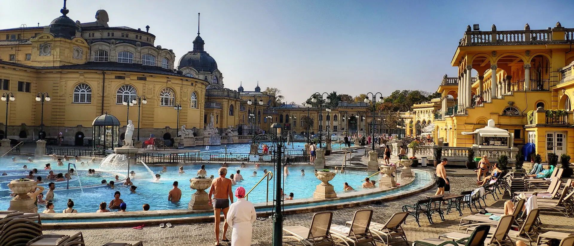 A sunny outdoor thermal bath complex with people swimming and lounging by the pool. Elegant historic buildings surround the area.