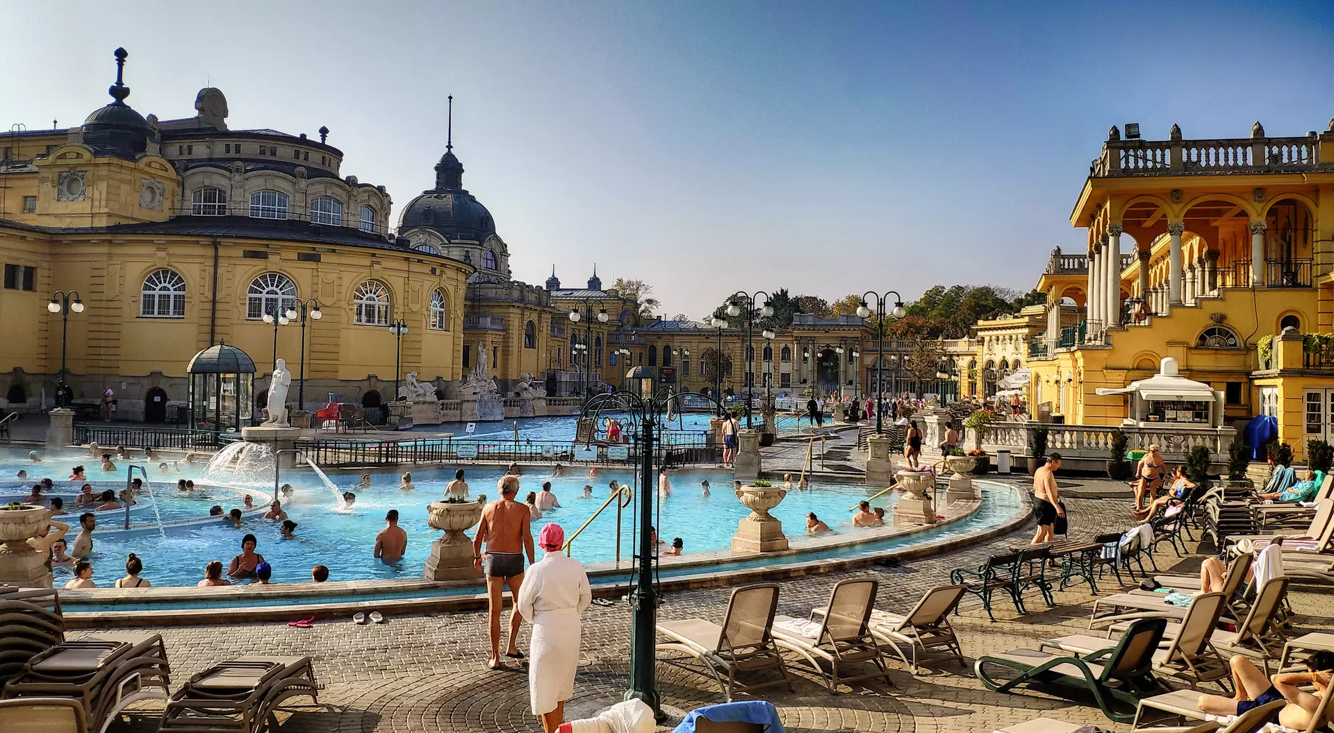 A sunny outdoor thermal bath complex with people swimming and lounging by the pool. Elegant historic buildings surround the area.