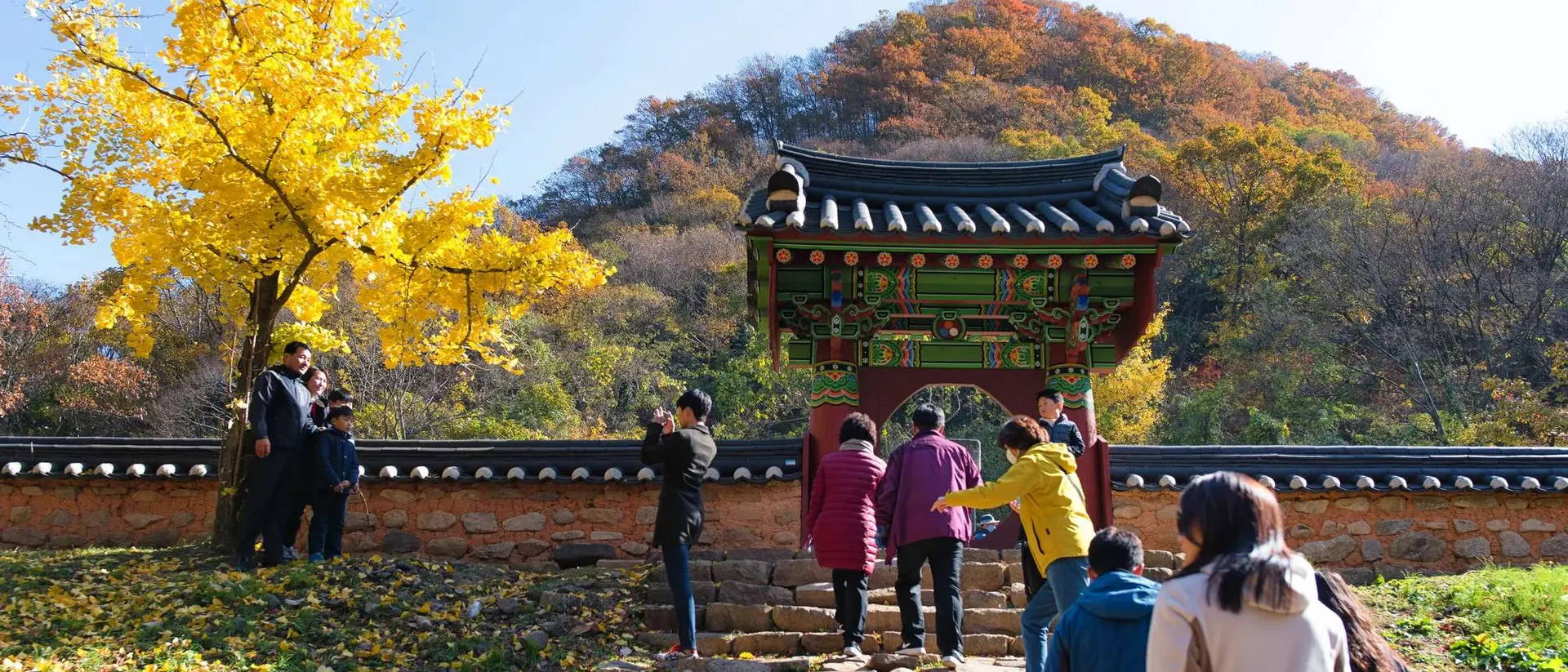 Visitors ascend stone steps toward a colorful traditional gate, surrounded by autumn trees and vibrant foliage.
