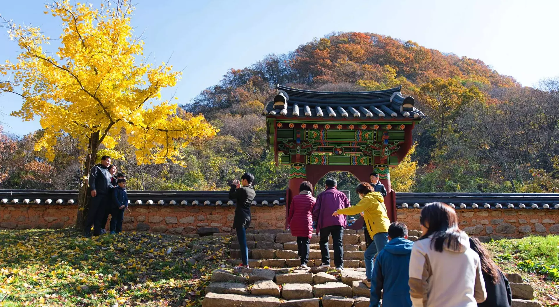 Visitors ascend stone steps toward a colorful traditional gate, surrounded by autumn trees and vibrant foliage.