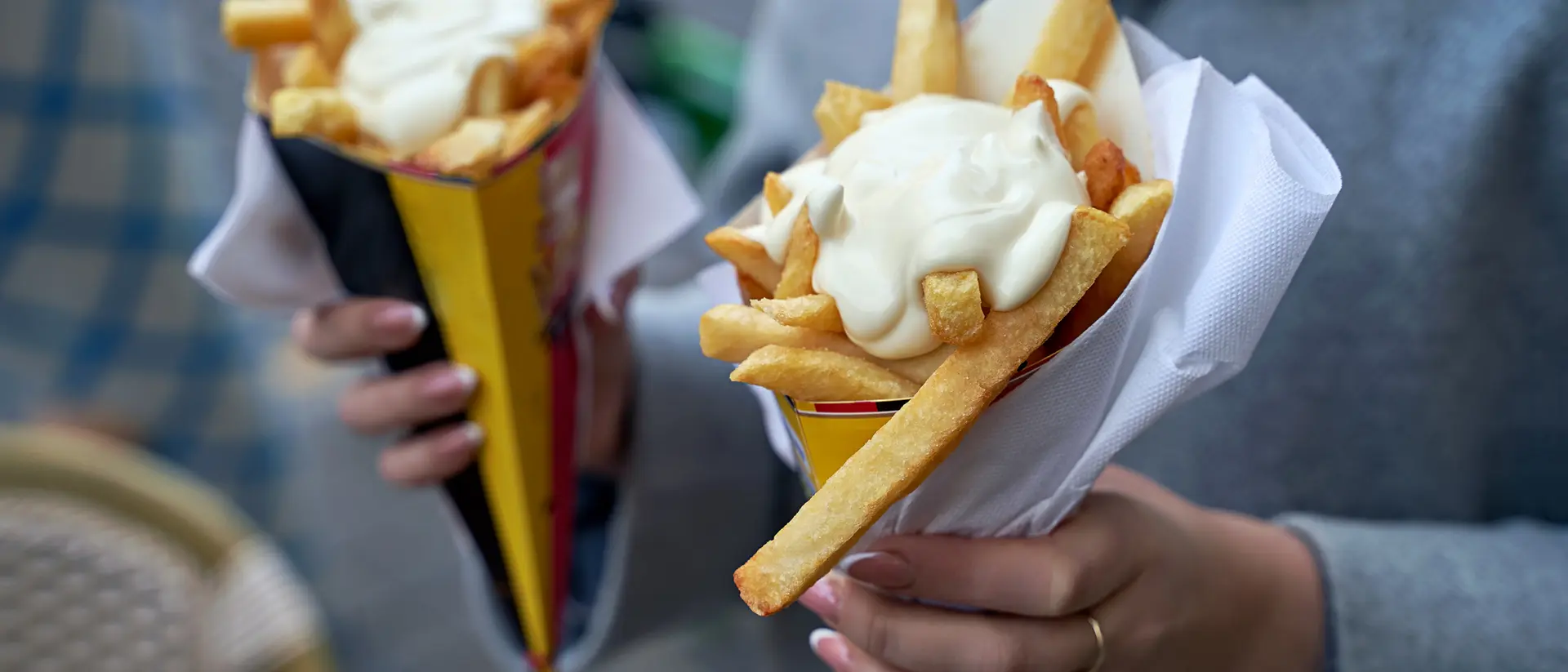 Two hands holding cones of fries topped with sauce, outdoors in a casual setting.