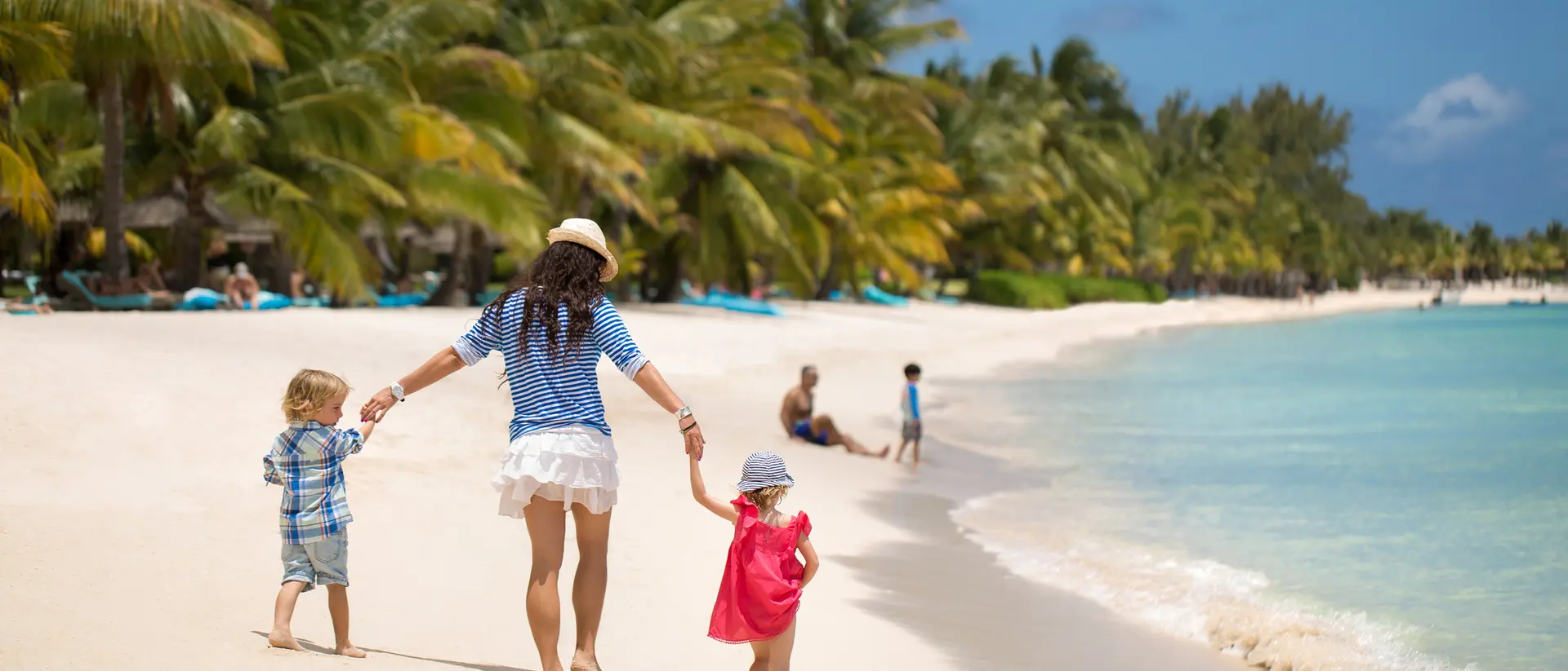 A woman holds hands with two children as they walk along a sunny beach, palm trees in the background, and gentle waves.