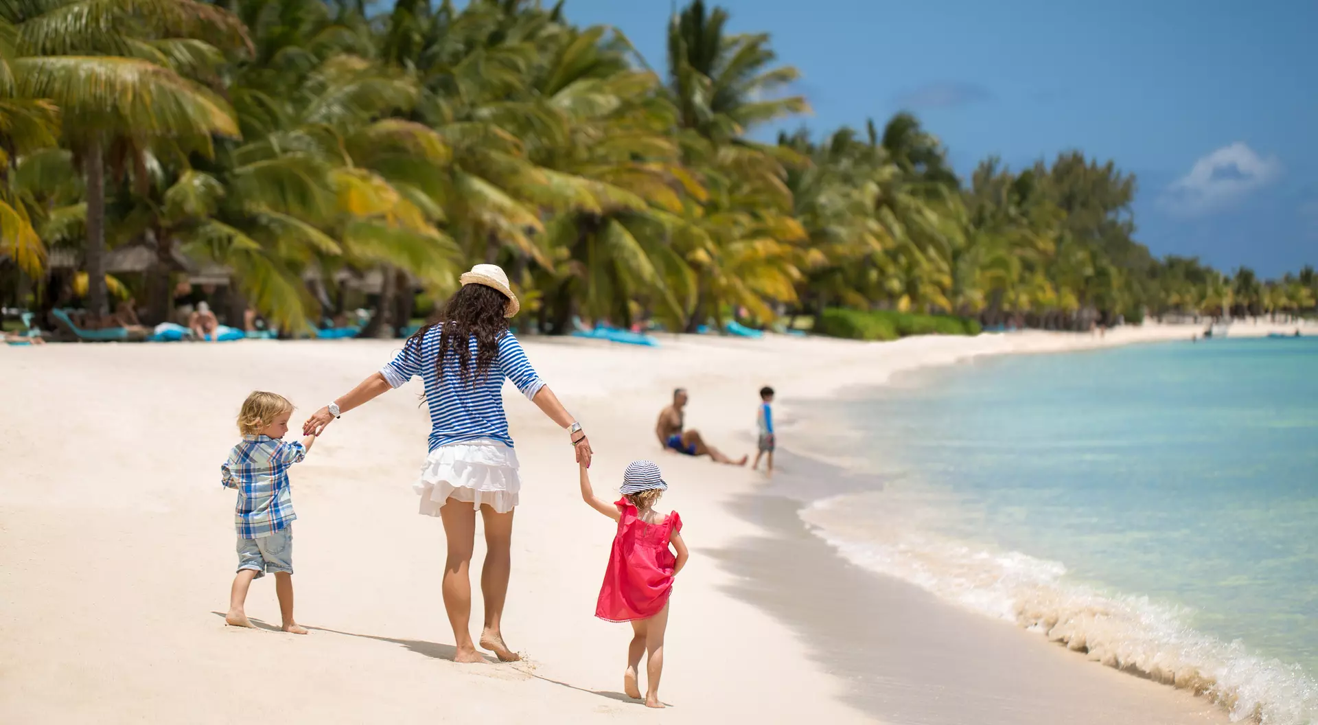 A woman holds hands with two children as they walk along a sunny beach, palm trees in the background, and gentle waves.
