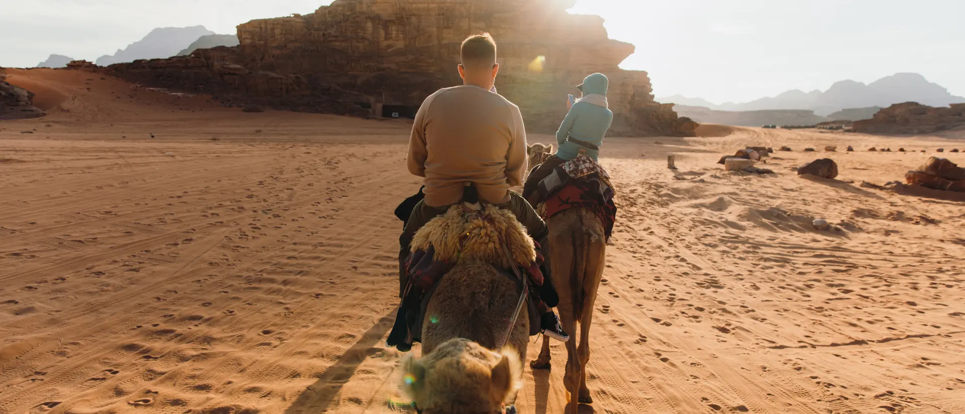 Two people riding camels through a sandy desert landscape, with mountains and sunlight in the background.