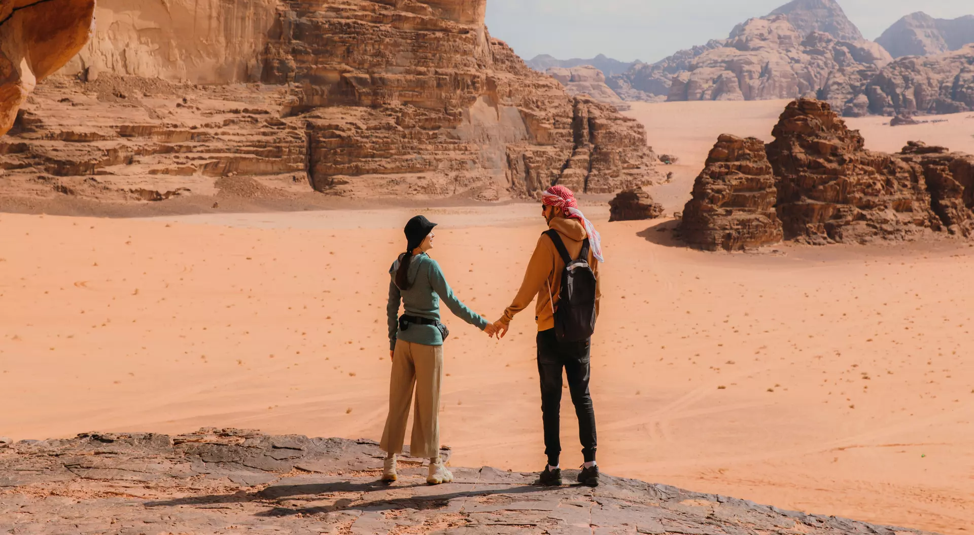 A couple holds hands in a desert landscape, surrounded by rocky formations and distant mountains.