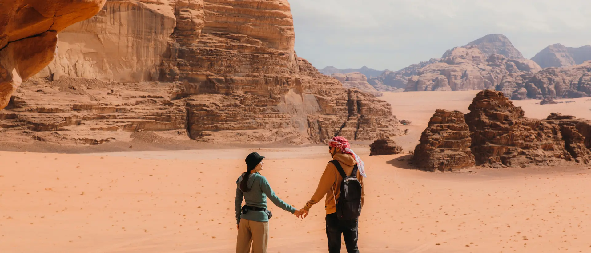 A couple holds hands in a desert landscape, surrounded by rocky formations and distant mountains.