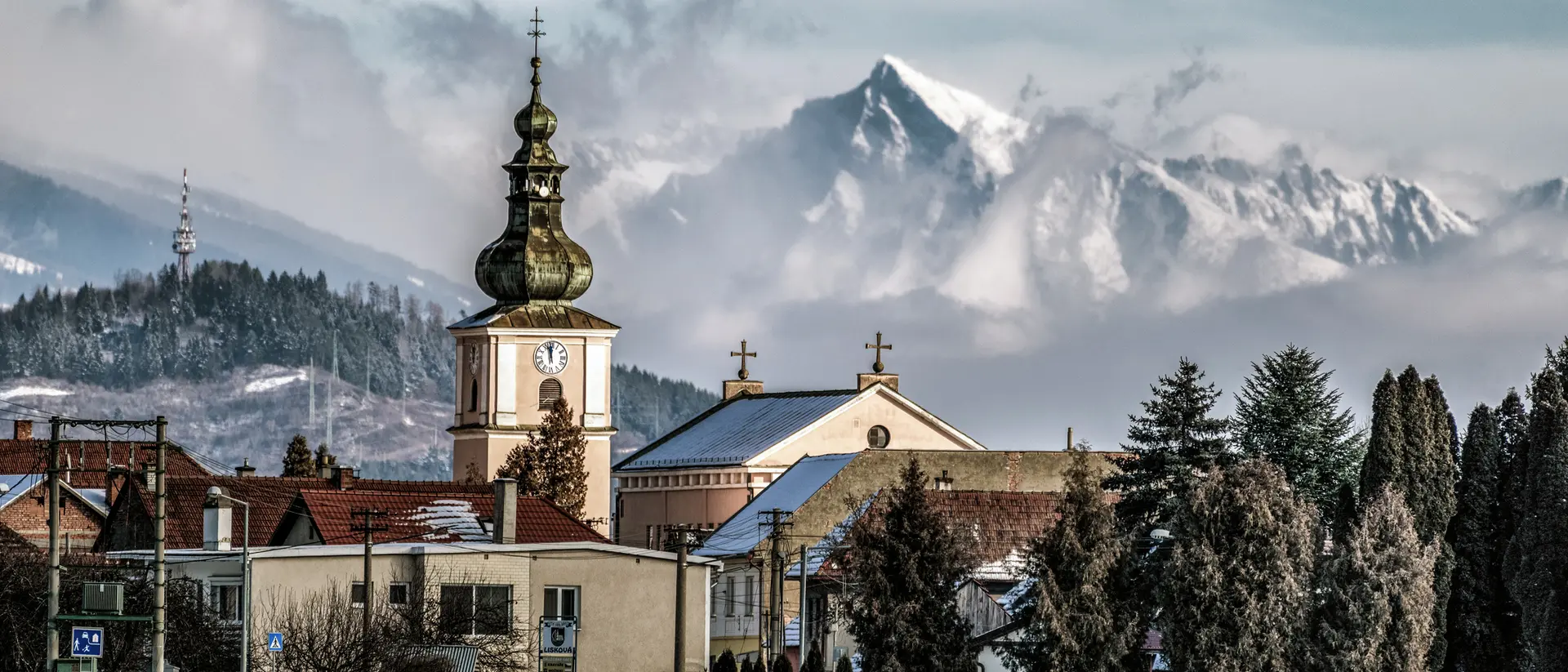 A snowy landscape with a church tower and mountains in the background, under a cloudy sky.
