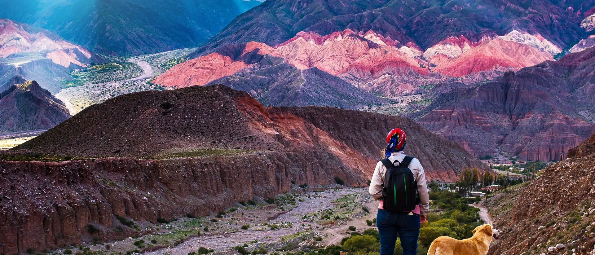 A person stands on a mountain path with a dog, overlooking colorful, layered hills and valleys in the background.