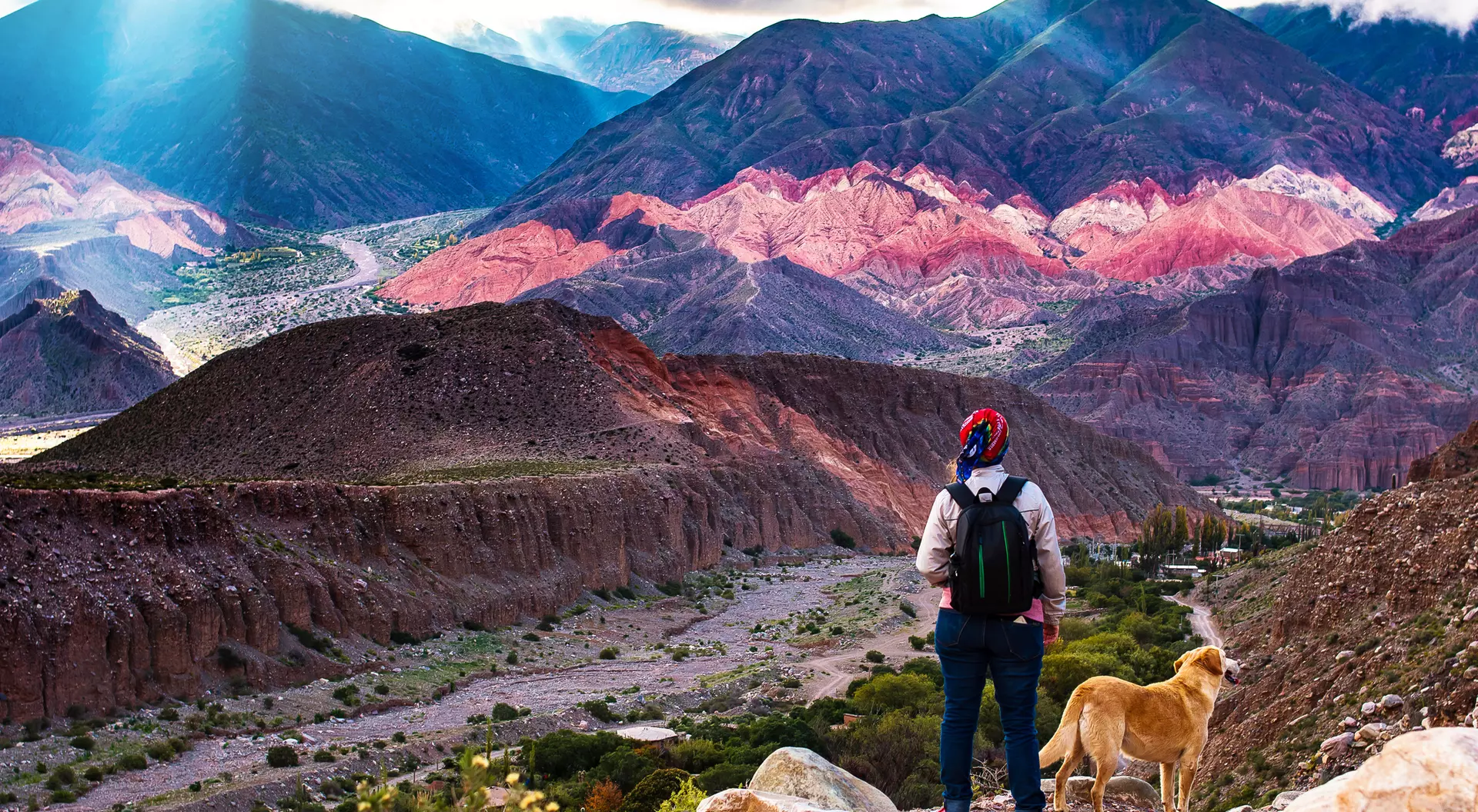 A person stands on a mountain path with a dog, overlooking colorful, layered hills and valleys in the background.