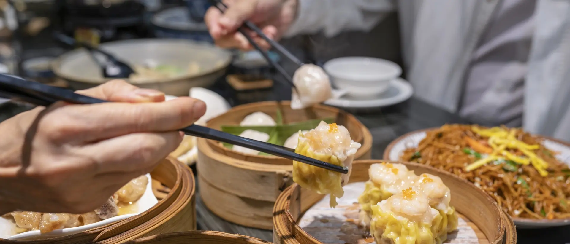Hands using chopsticks to pick up dim sum from bamboo steamers on a table with noodles and other dishes in the background.
