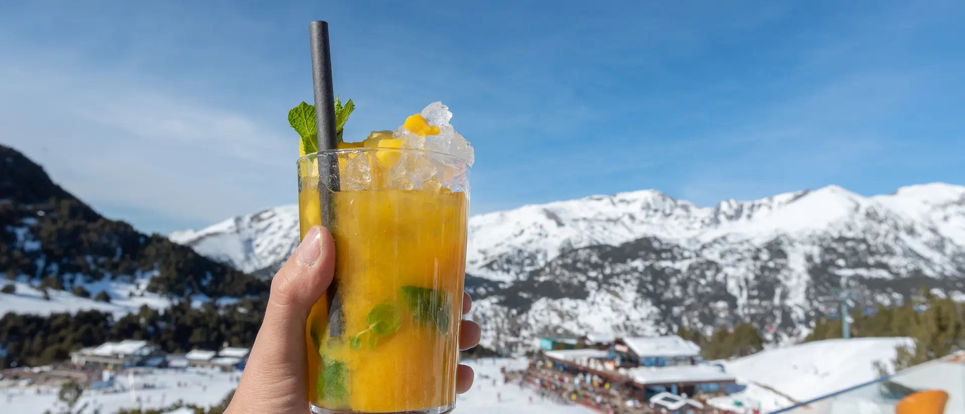 A hand holds a glass of orange cocktail with mint, set against a snowy mountain backdrop under a clear blue sky.