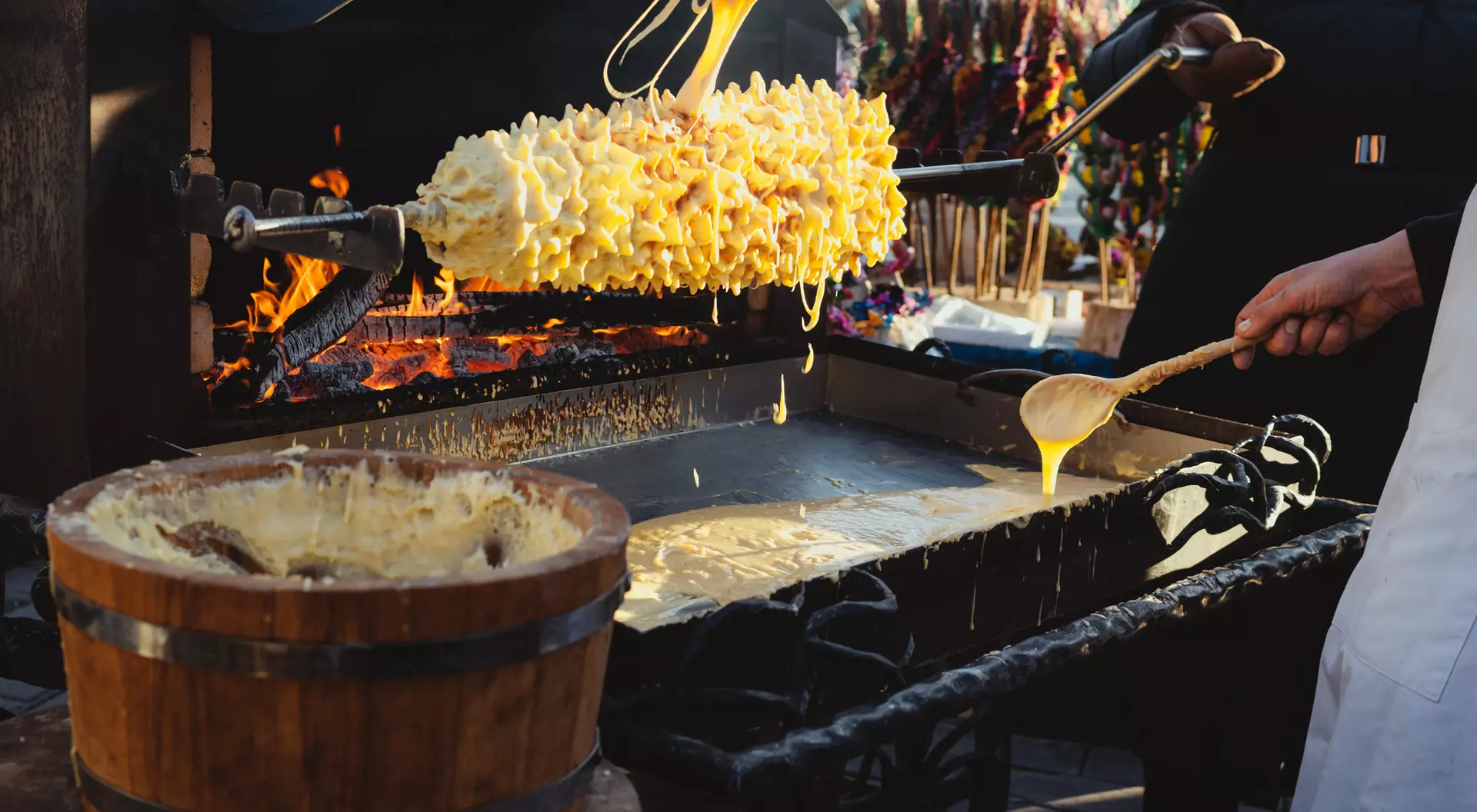A person holds a large, cheese-covered food on a skewer over a hot grill, with melted cheese dripping into a bowl below.