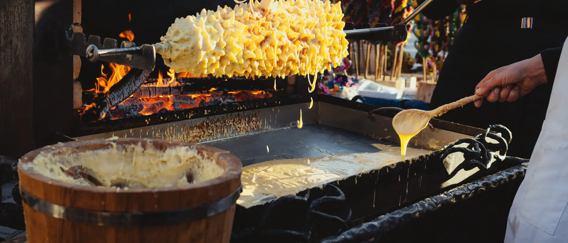A person holds a large, cheese-covered food on a skewer over a hot grill, with melted cheese dripping into a bowl below.