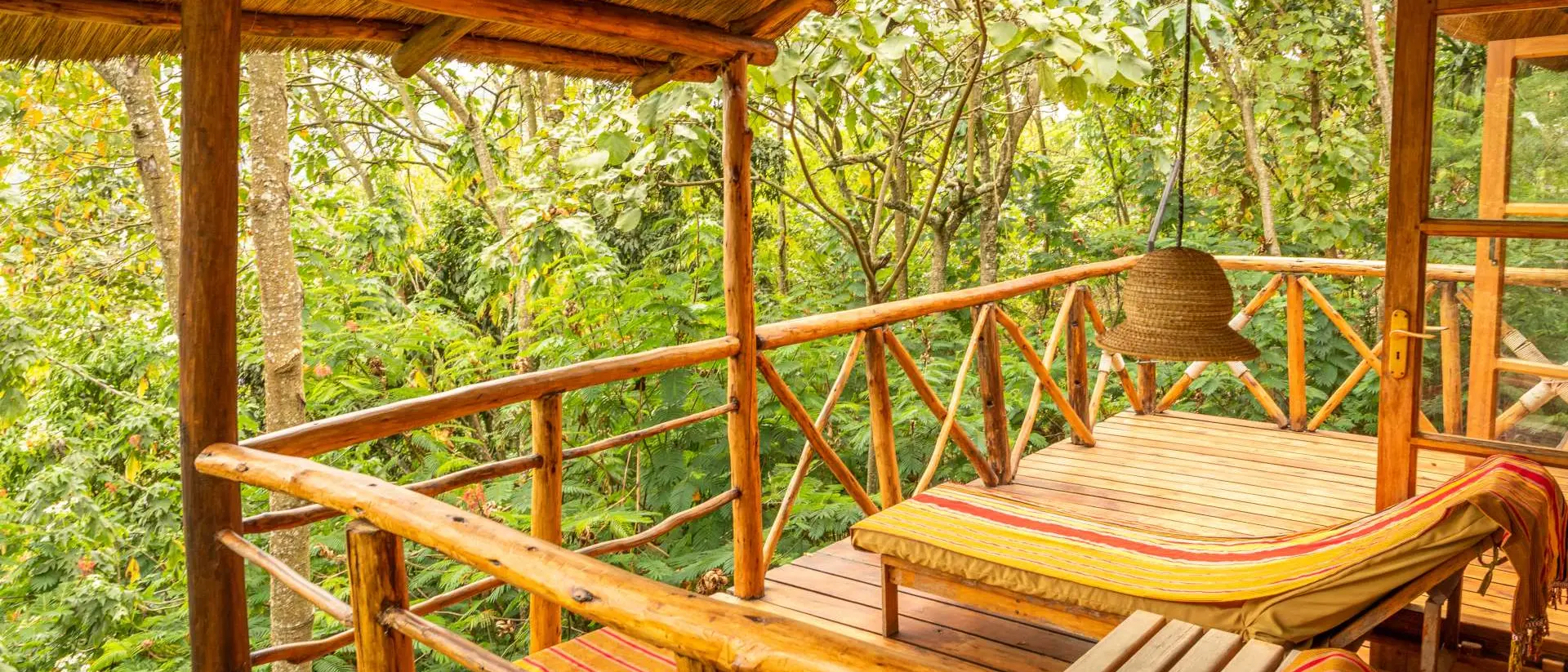 Cozy wooden balcony with striped loungers, surrounded by lush greenery and a hanging light fixture.