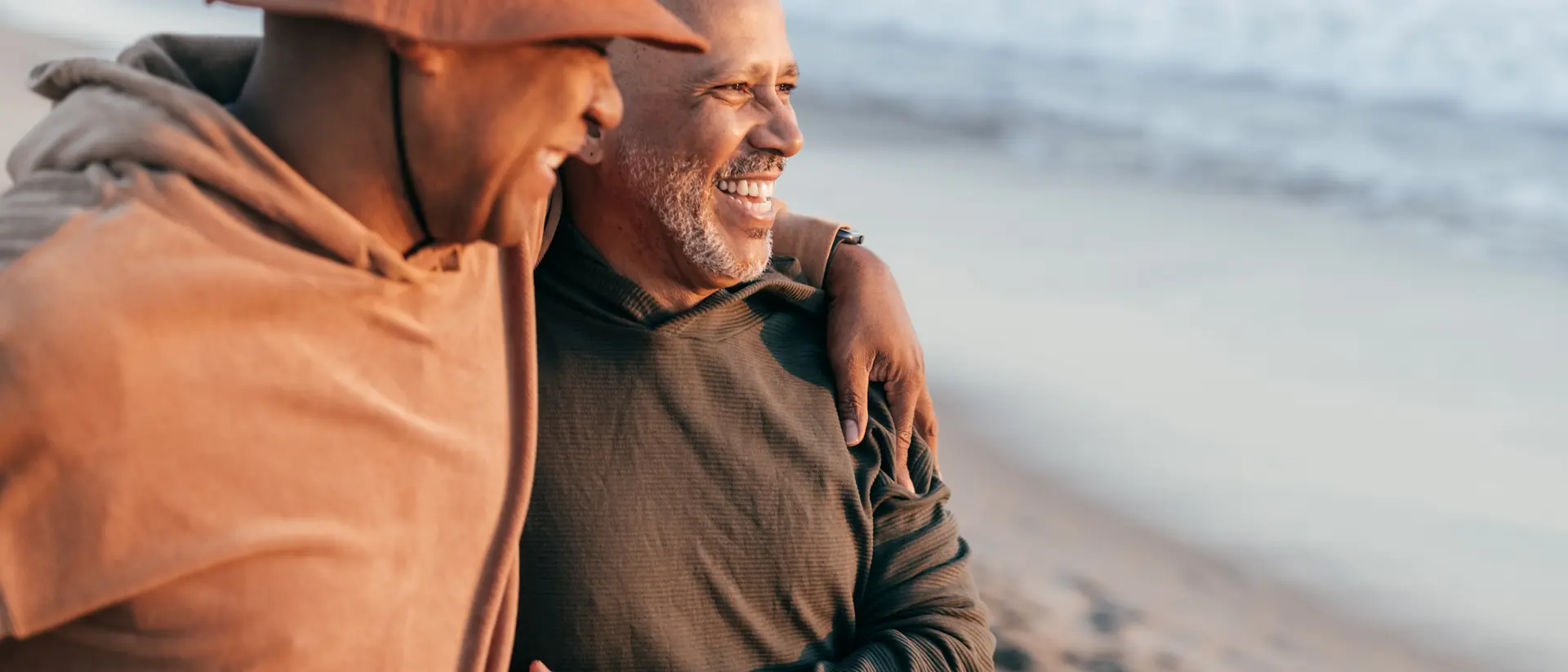 Two men smile and walk together on a beach, enjoying the moment with the ocean in the background.
