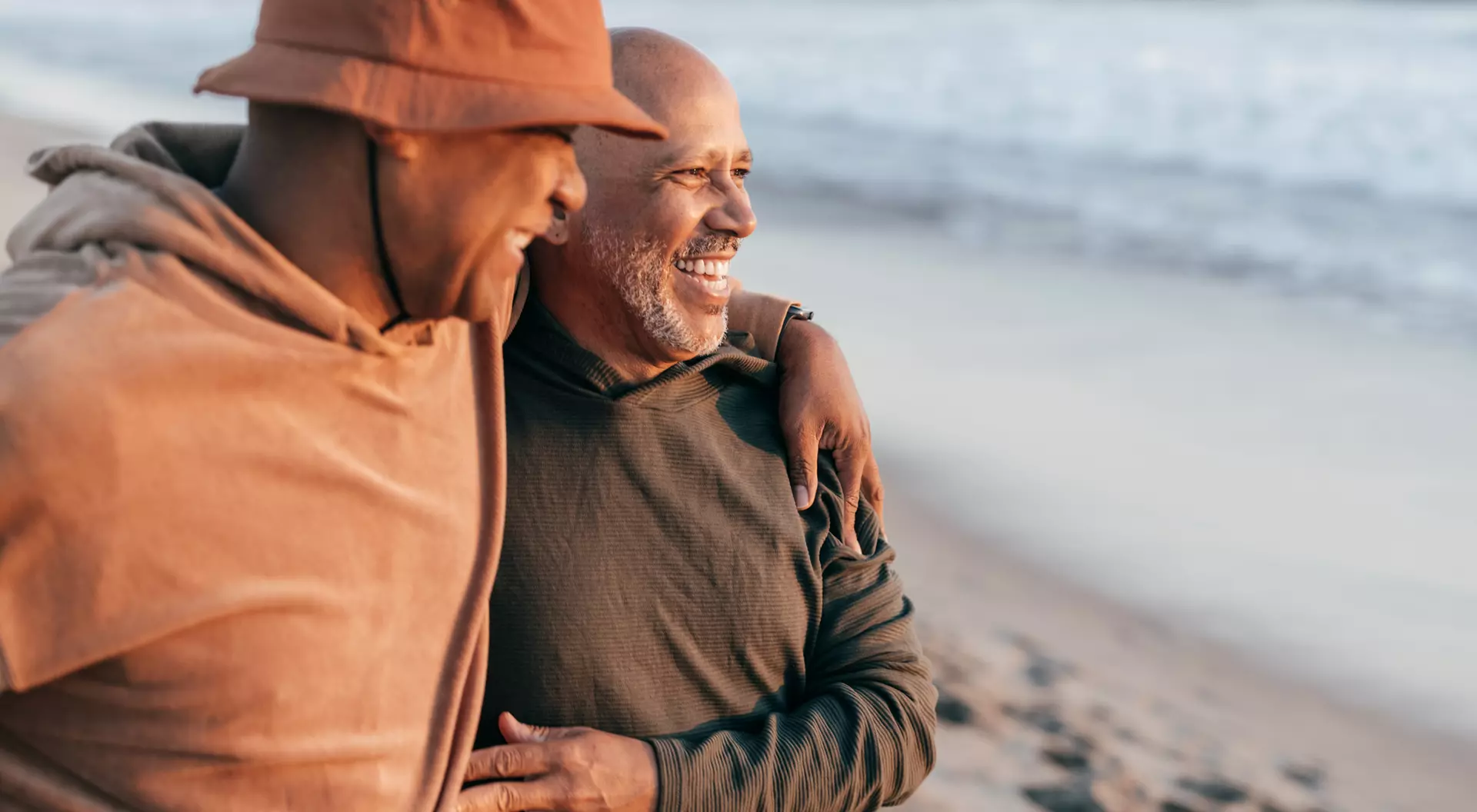 Two men smile and walk together on a beach, enjoying the moment with the ocean in the background.