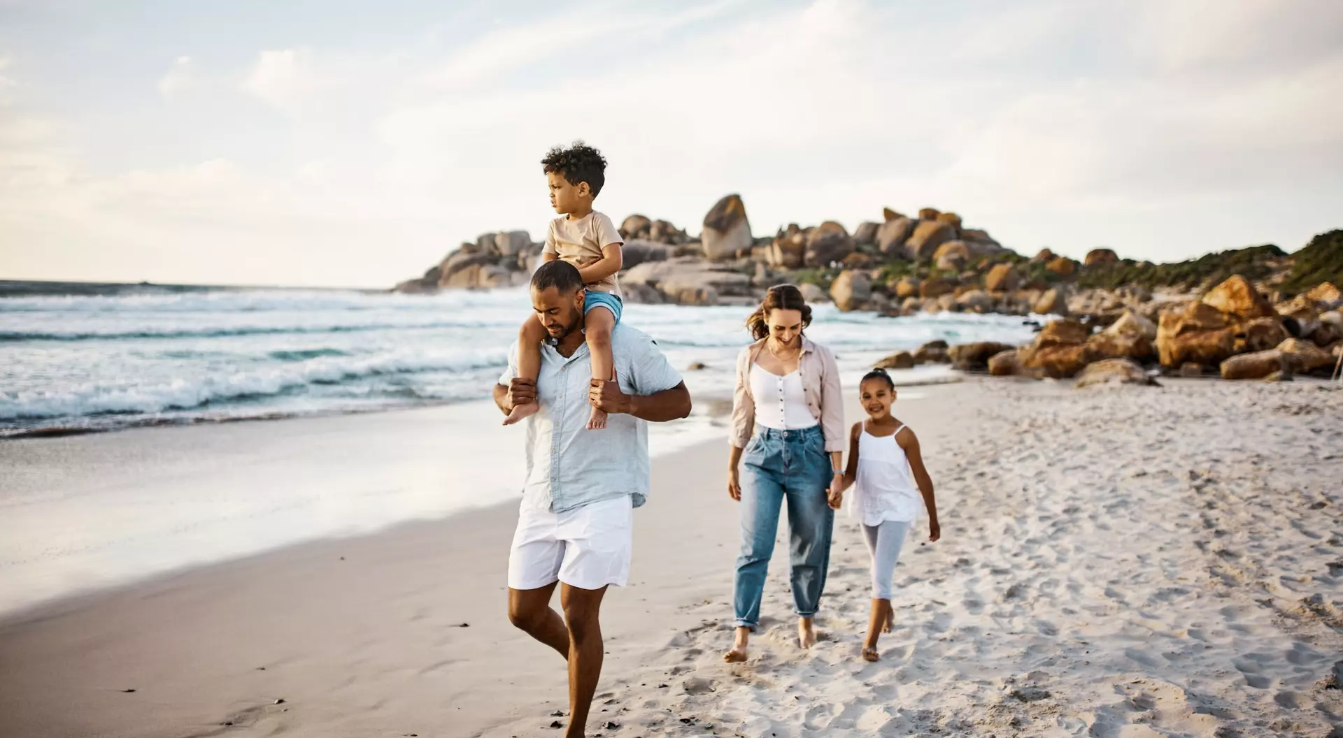 A family walks along a beach, with a man carrying a child on his shoulders and two girls walking beside them.