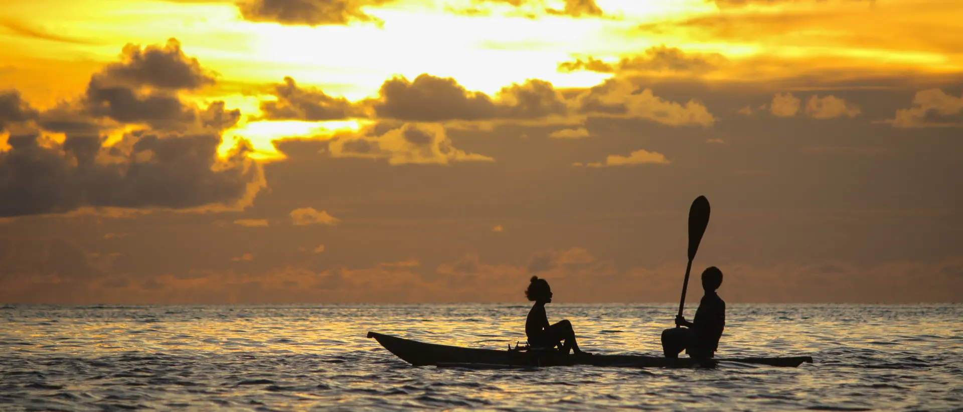 Two people sit in a kayak at sunset, silhouetted against a colorful sky with clouds reflecting in the water.
